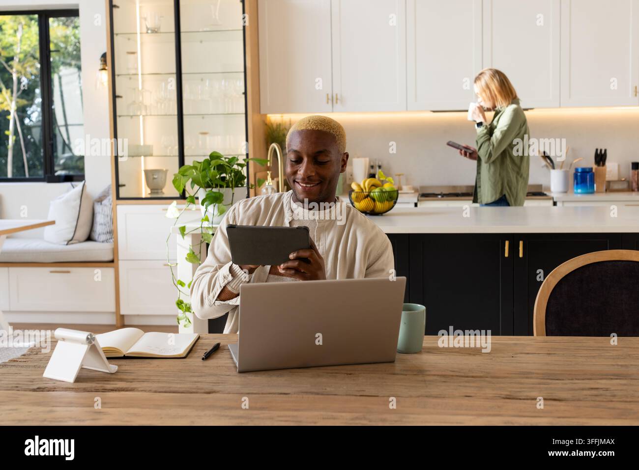 Diverse couple sitting at table in kitchen, man using tablet with laptop, woman holding smartphone Stock Photo