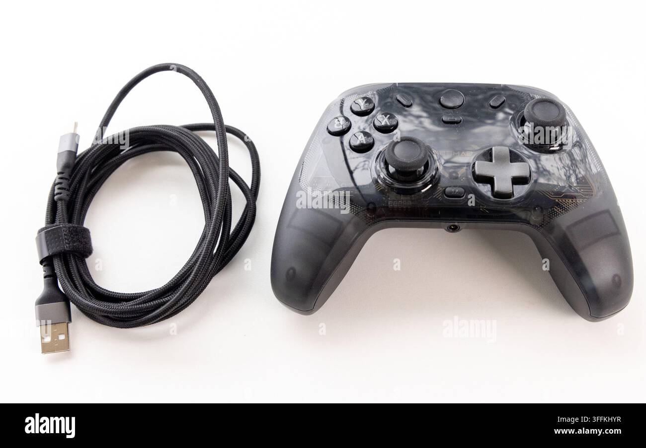 A transparent black game controller and coiled USB cable rest on white Stock Photo