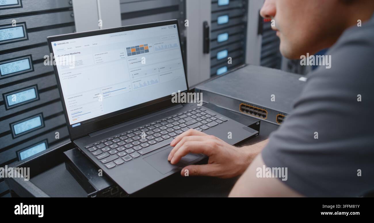IT Administrator Uses Laptop Beside Network Switch to Navigate Server, Network and Security Logs, Comparing Software Metrics with Hardware Status Thro Stock Photo