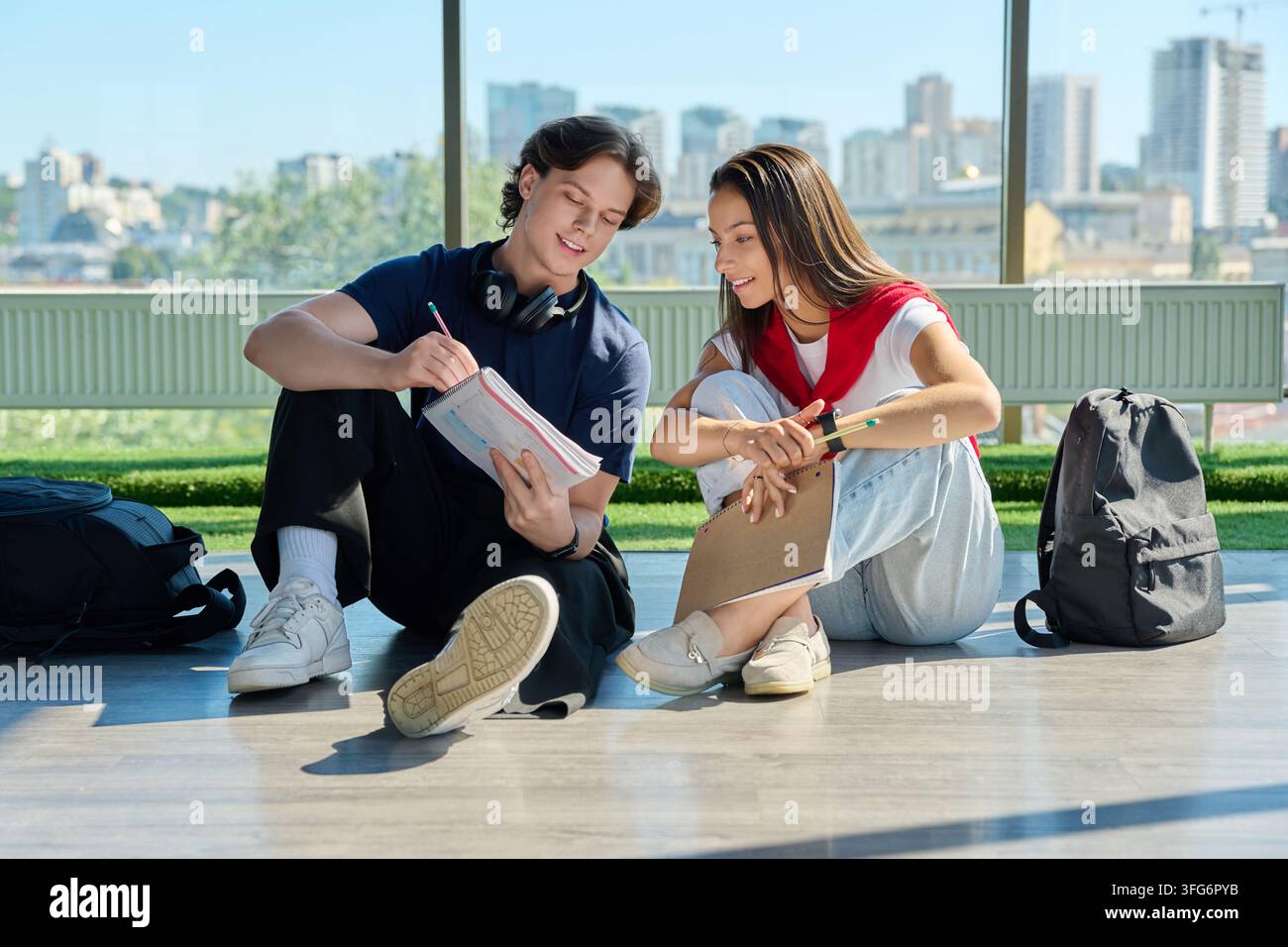 Two young students studying together with notebooks sitting on floor near big window Stock Photo