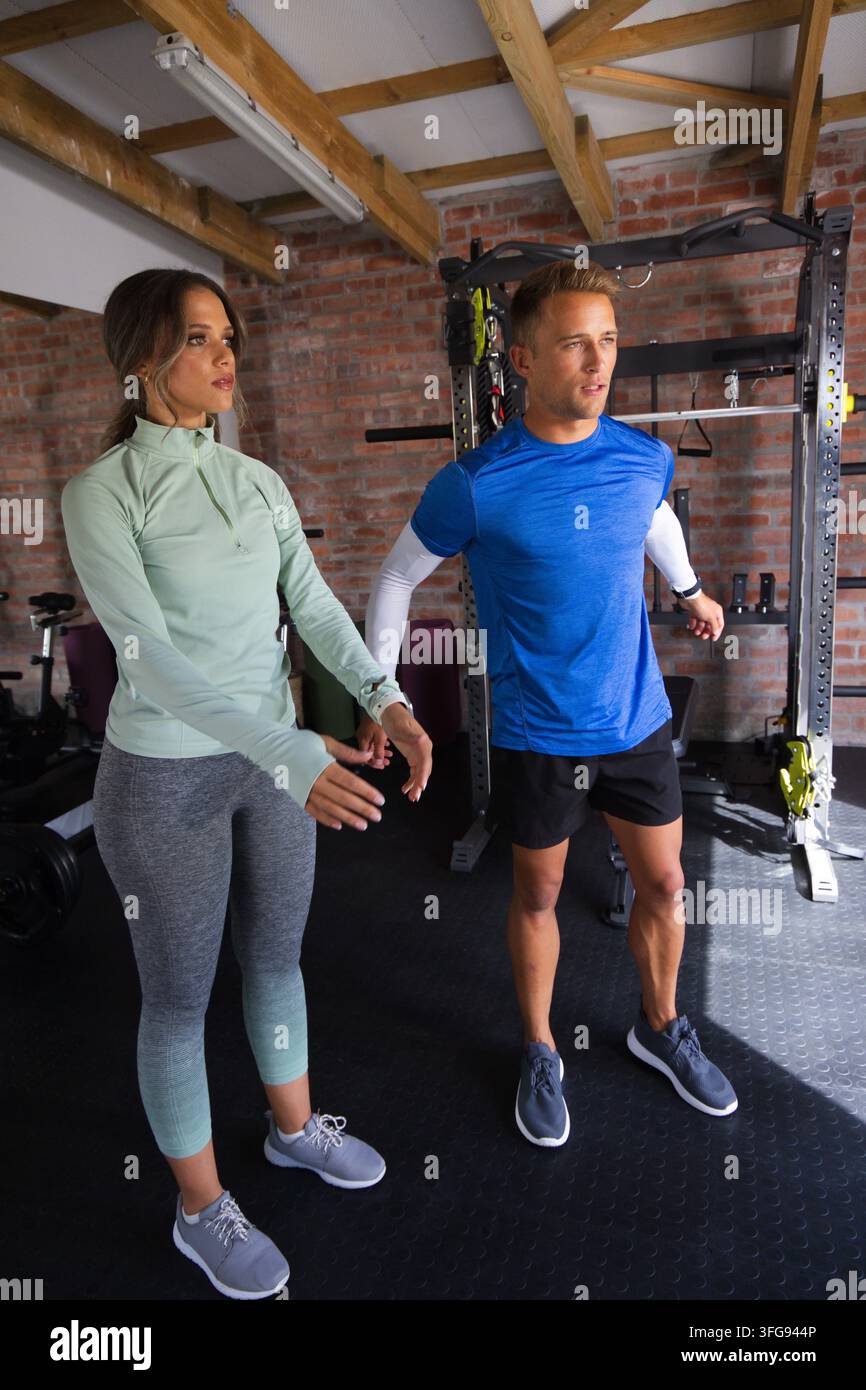 Diverse couple doing warm-up stretches in garage-style gym wearing athletic clothes near power rack Stock Photo