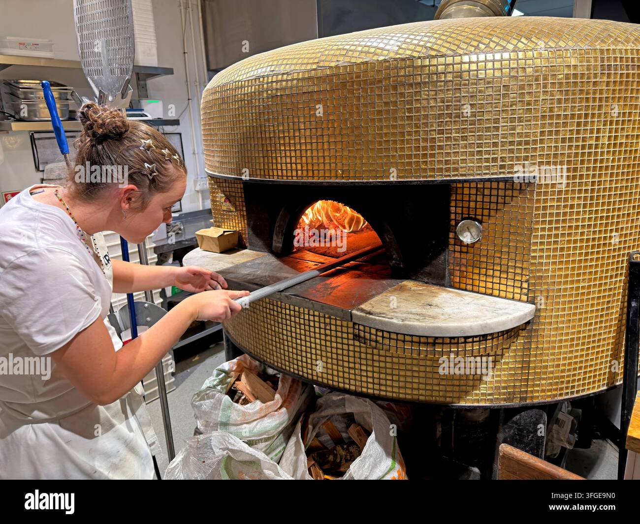 Pizza chef tending a wood-fired oven with a long-handled peel in a traditional Italian pizzeria restaurant kitchen. Stock Photo