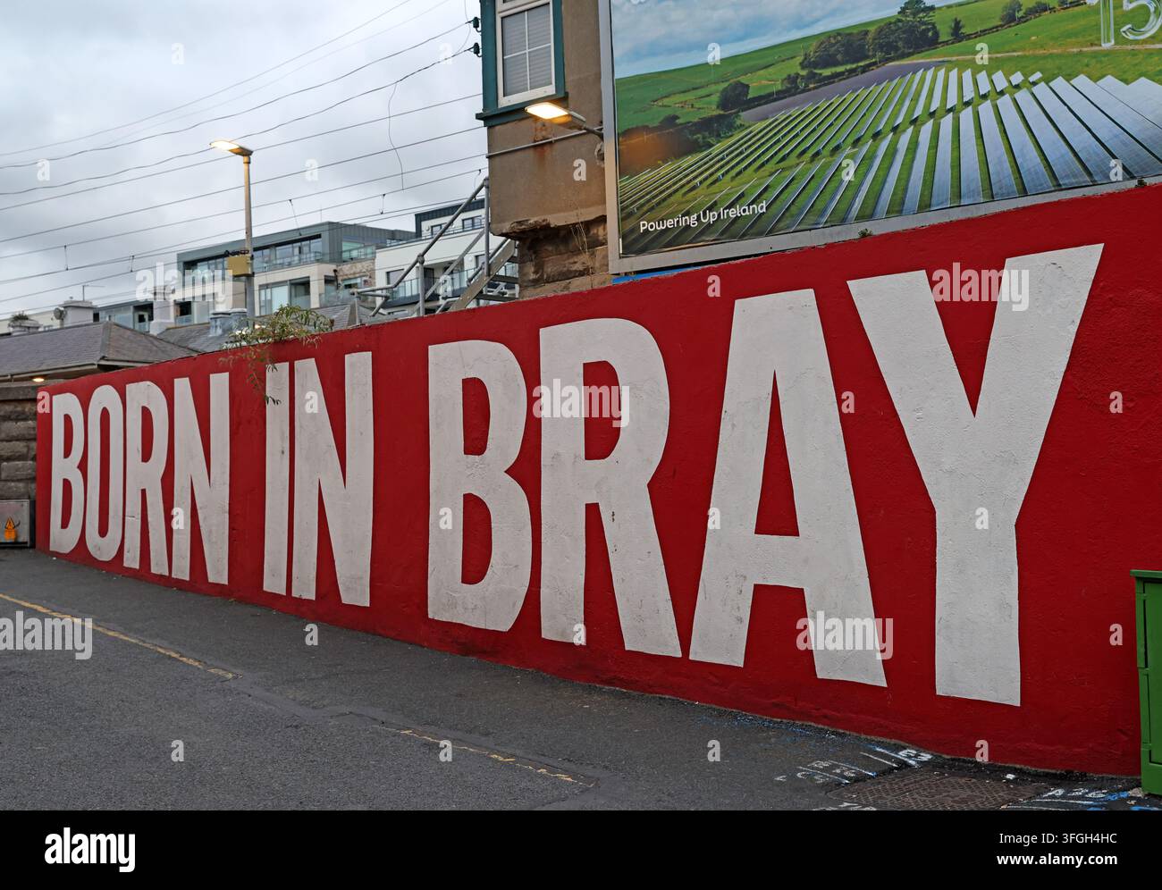 Bold red “Born in Bray” mural beside the DART railway in Bray, County Wicklow, Ireland, with a solar-energy billboard overlooking the line. Stock Photo