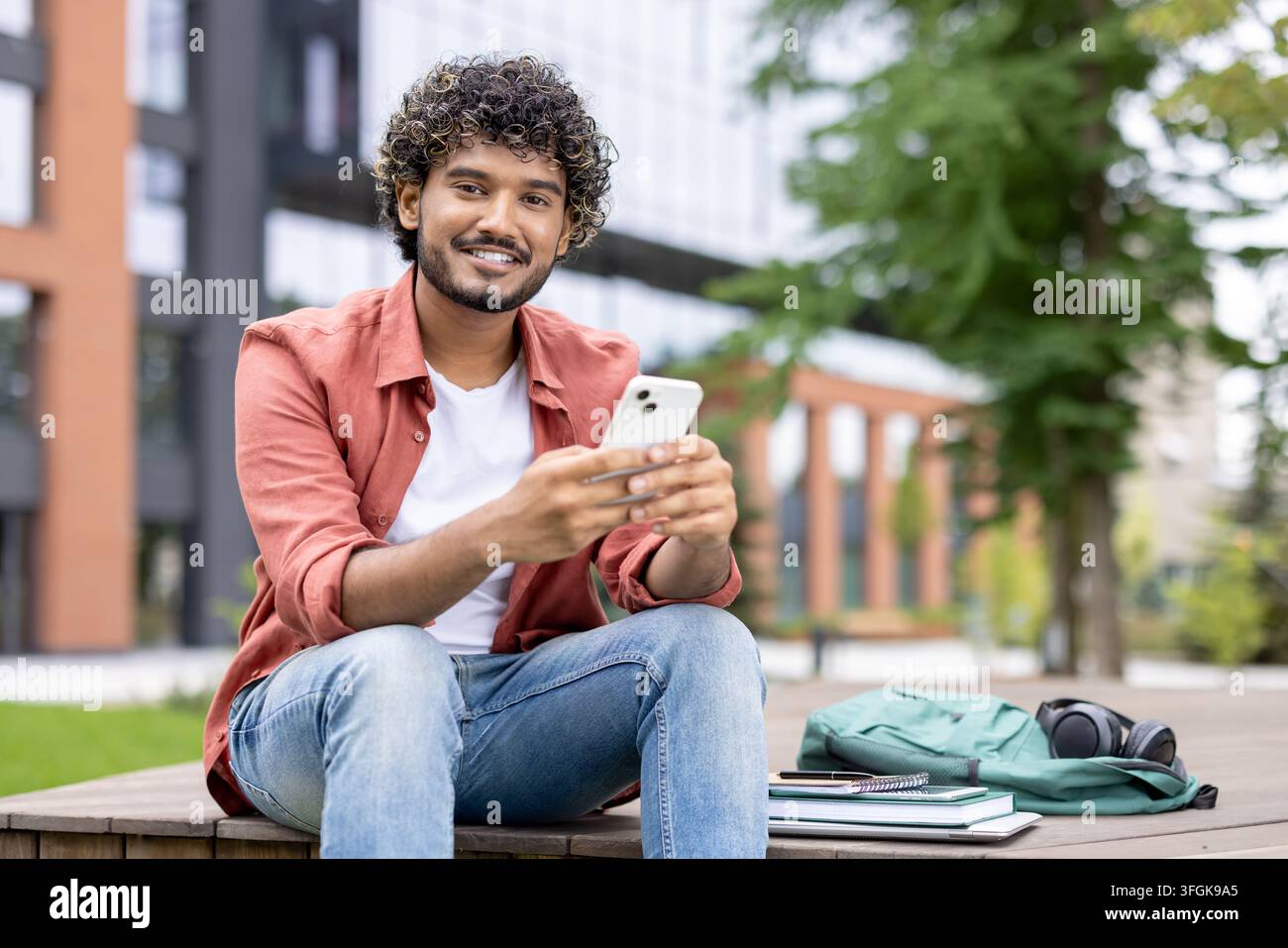 Smiling student drinking takeaway coffee and using smartphone during study break on campus grounds. Stock Photo