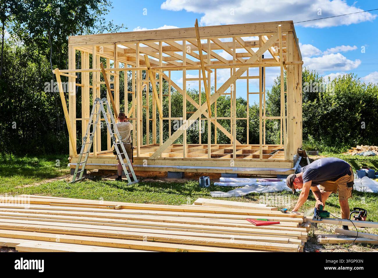 Construction crew assembles wooden frame with circular saw and lumber while one worker cuts boards and second secures bracing inside structure. Stock Photo