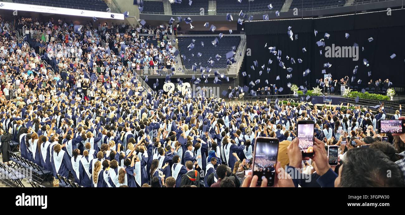 Graduating high school students throw their caps into the air during a graduation ceremony as families and guests celebrate from the stands in a large - Smartphone Captured Stock Image