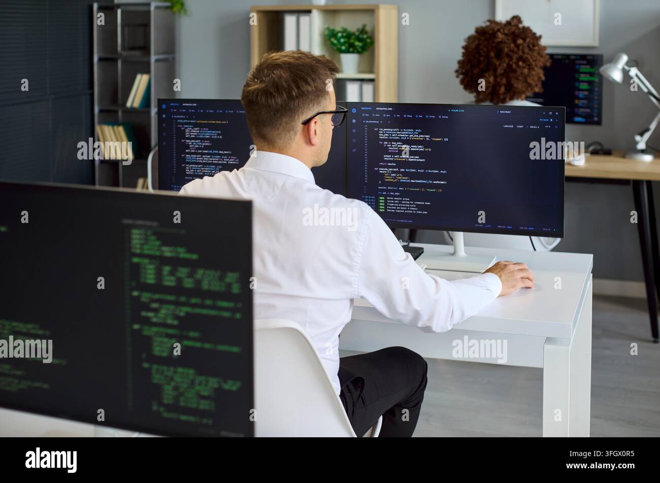 Rear view of developer coding at dual monitors in contemporary tech office Stock Photo