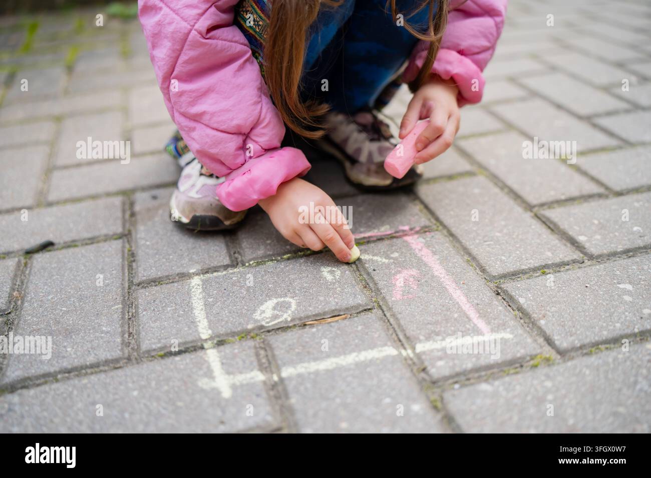 Child drawing with chalk on paving stones while holding another chalk stick, illustrating hands on creative play and the practice of early drawing ski Stock Photo