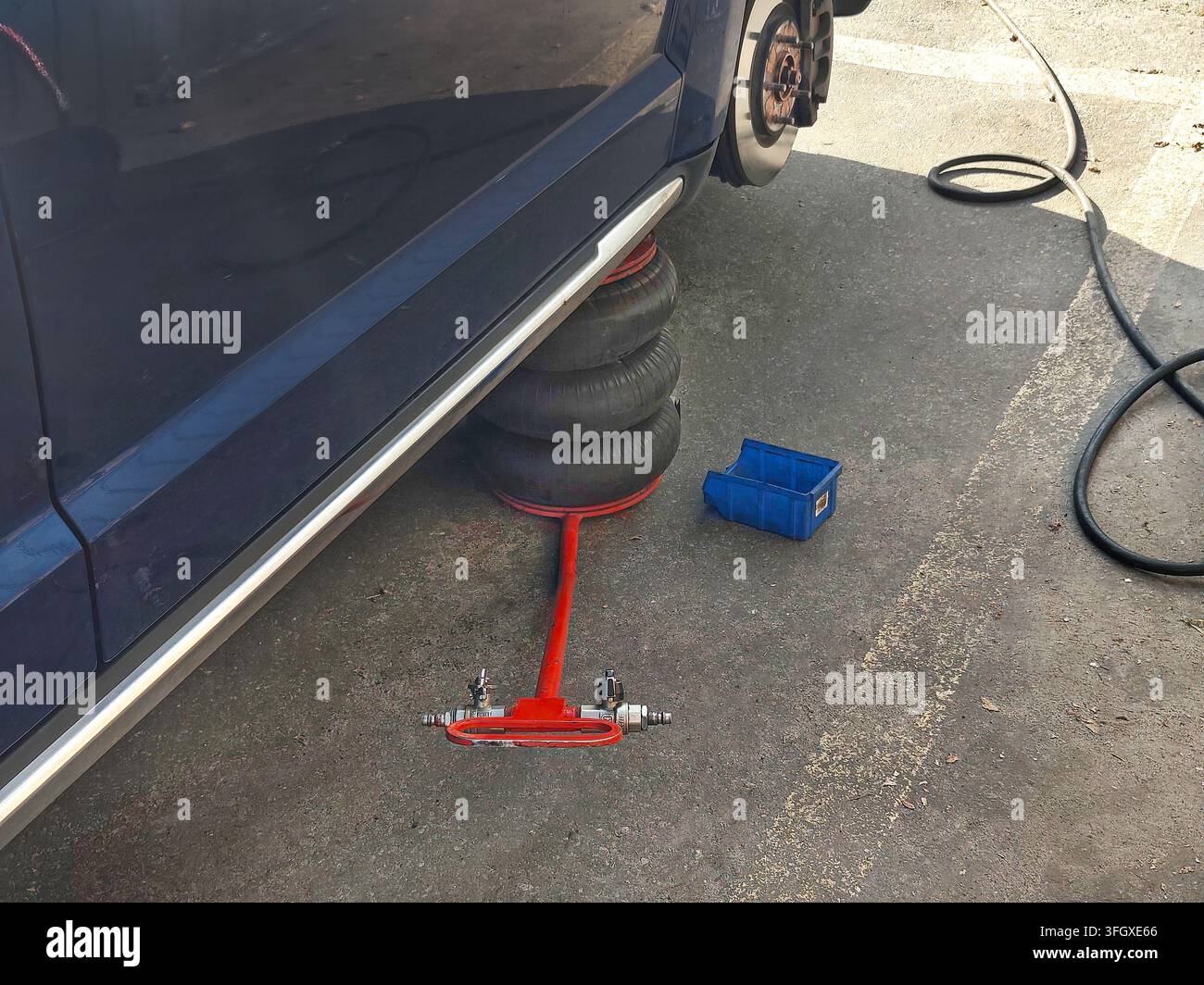 Blue car lifted with a red hydraulic jack and stacked tires underneath, with a blue plastic crate and black hose on the concrete ground beside it. Stock Photo