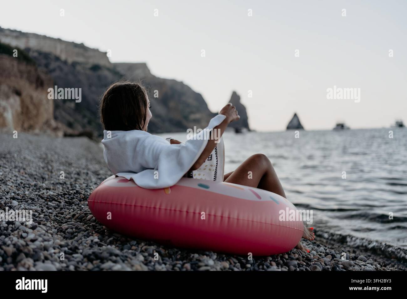 Woman, beach, sea. Young woman on inflatable ring relaxes on pebble beach, enjoying tranquil water, cliffs at dusk. Stock Photo