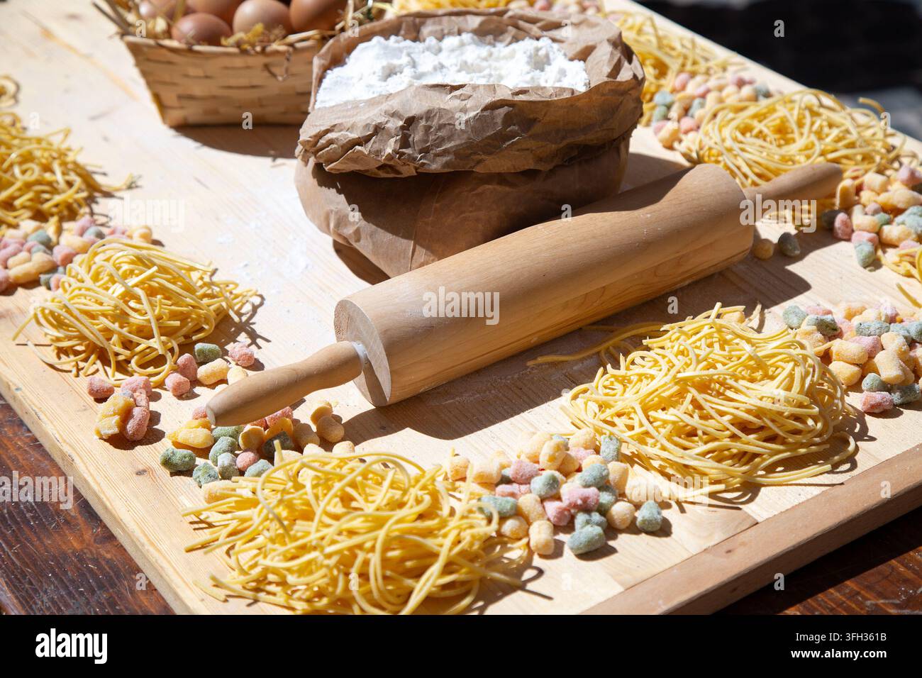 Homemade pasta ingredients with flour bag, wooden rolling pin, egg basket, and dried noodle nests arranged on rustic board in bright natural kitchen Stock Photo