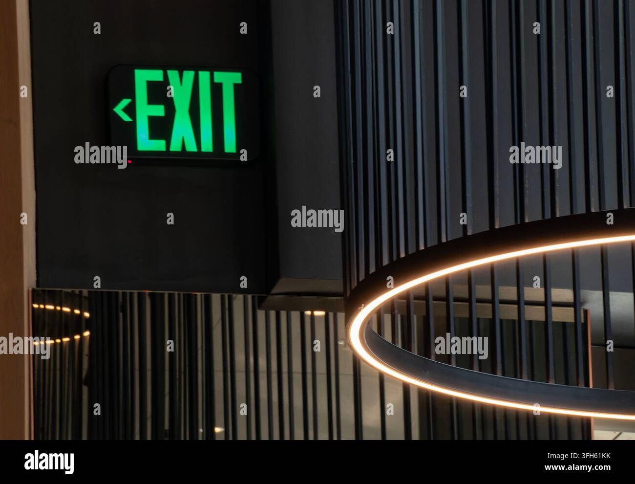 An illuminated green "EXIT" sign mounted on a dark wall next to a modern sloped ceiling fixture Stock Photo