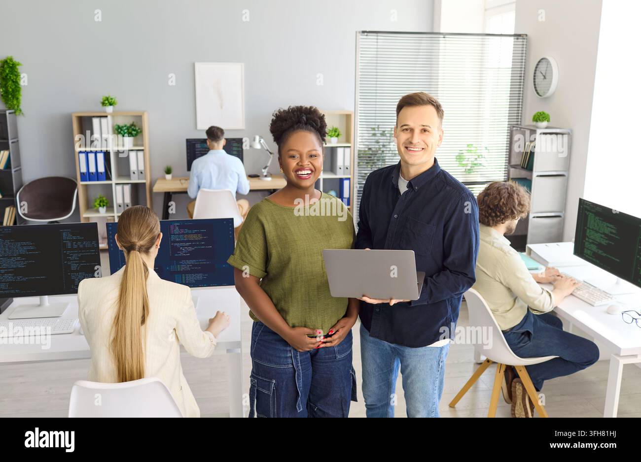 Smiling coworkers with laptop in bright office as programmers code at workstations Stock Photo