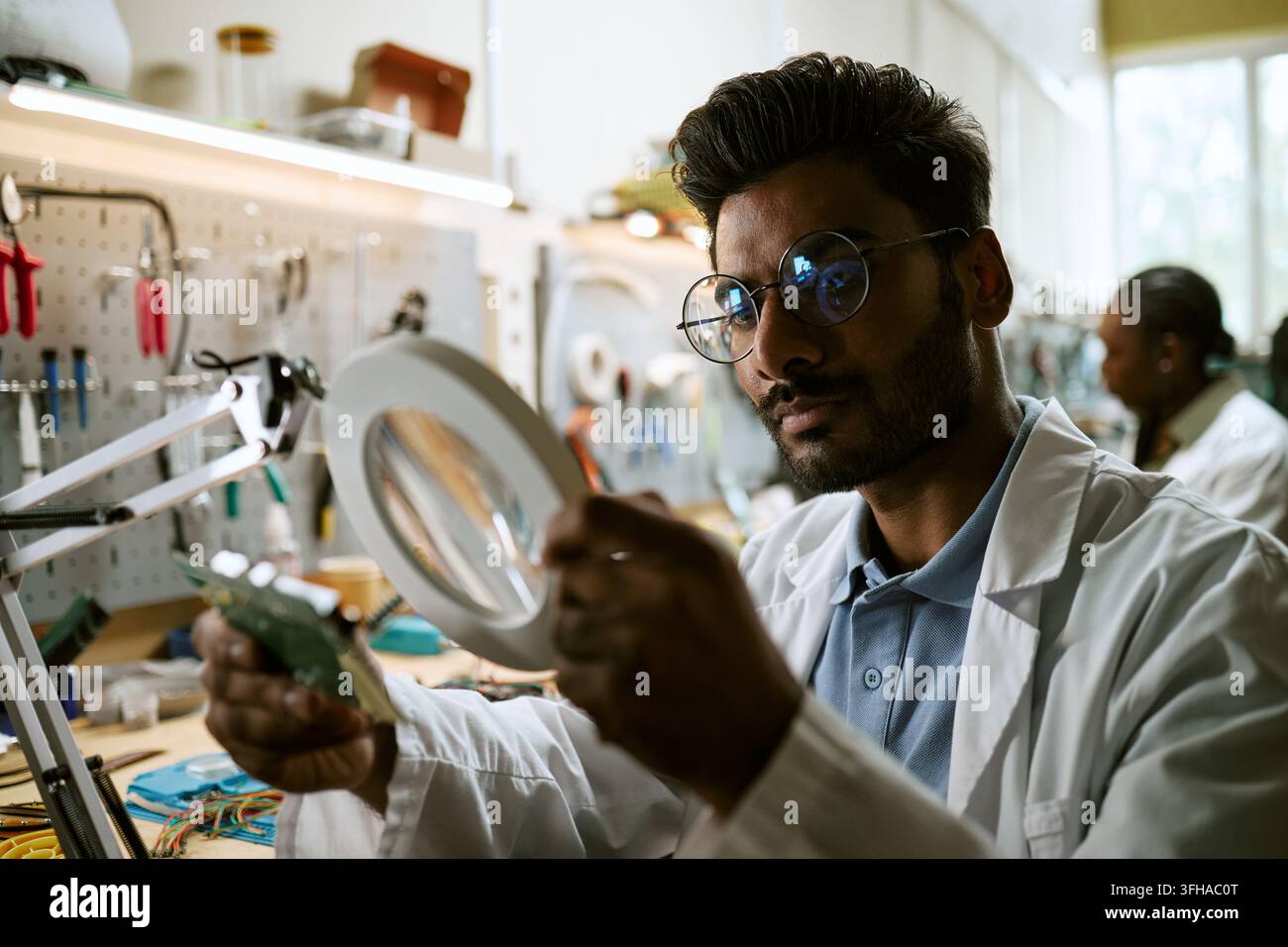 Indian Engineer Inspecting Circuit Board with Magnifier in Tech Lab Stock Photo