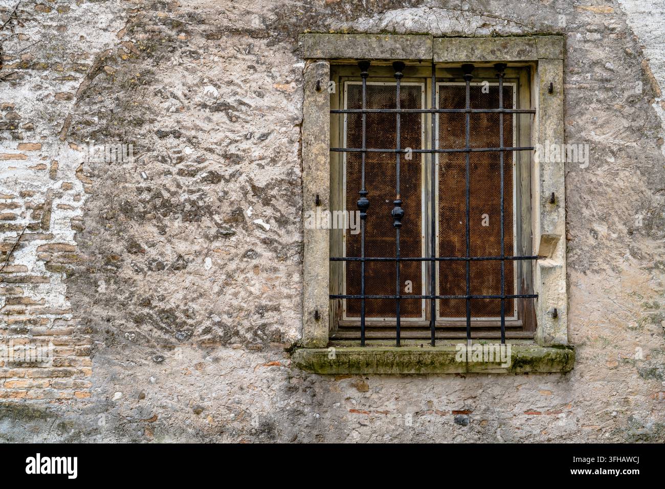 Old window protected by iron bars and framed in stone on a weathered masonry wall, showing aged plaster, exposed brick and traditional architectural d Stock Photo