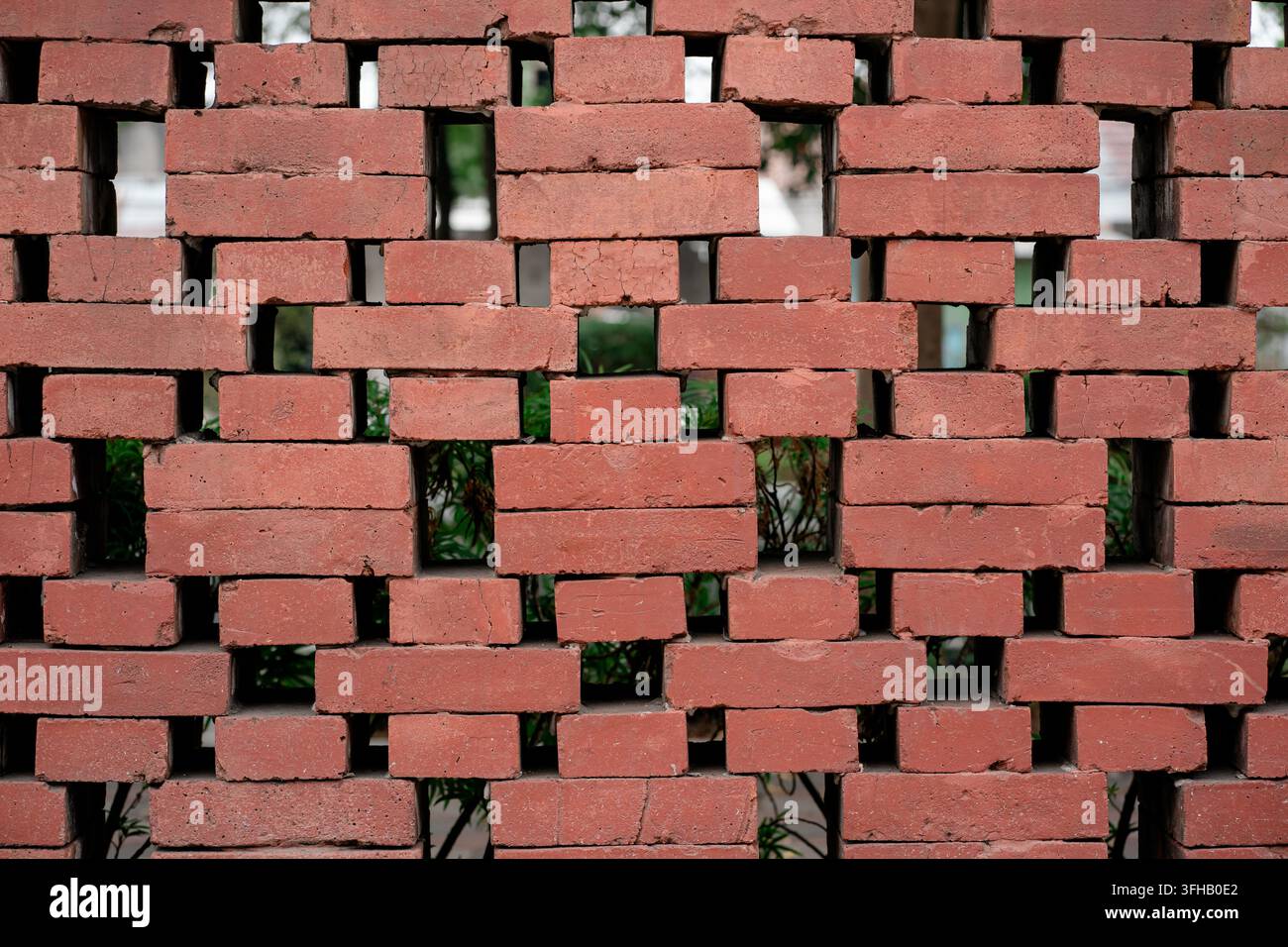 Close up of a decorative red brick wall with missing blocks creating a geometric ventilation pattern with green foliage behind. Stock Photo