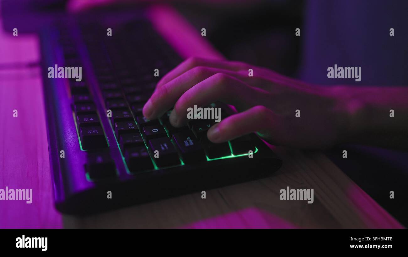 Young hispanic man typing with hands on rgb backlit gaming keyboard in studio at wooden desk under purple neon lighting; focused immersion. Stock Photo