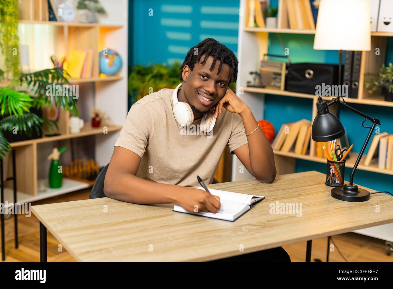 African American young man on home table writes notes in notebook with pen for study planning at home. Black guy on home table reviews written task list with happy focused mood in living room interior Stock Photo