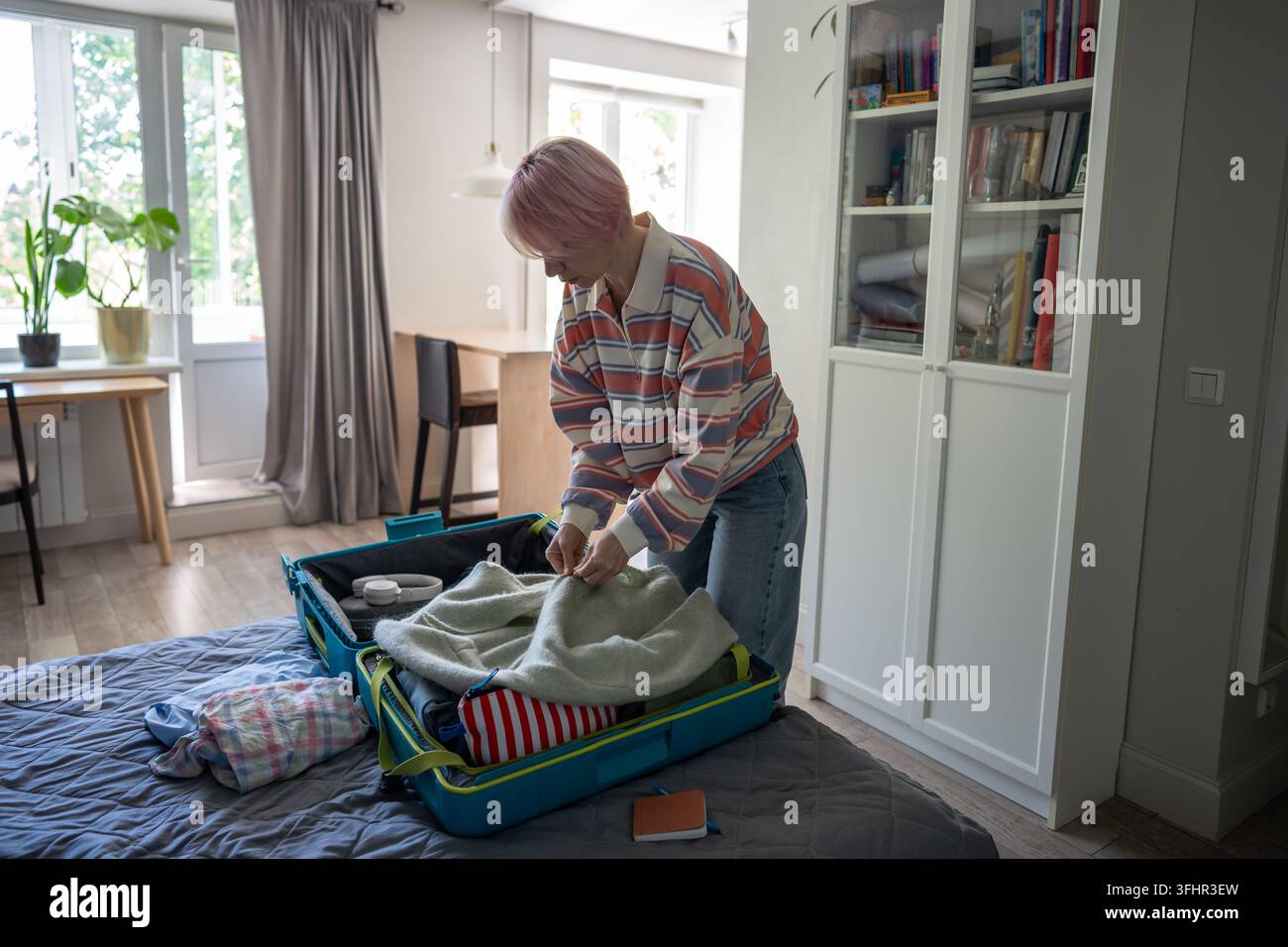 Woman thoughtfully preparing clothes for traveling on holiday and folding sweater in suitcase. Stock Photo