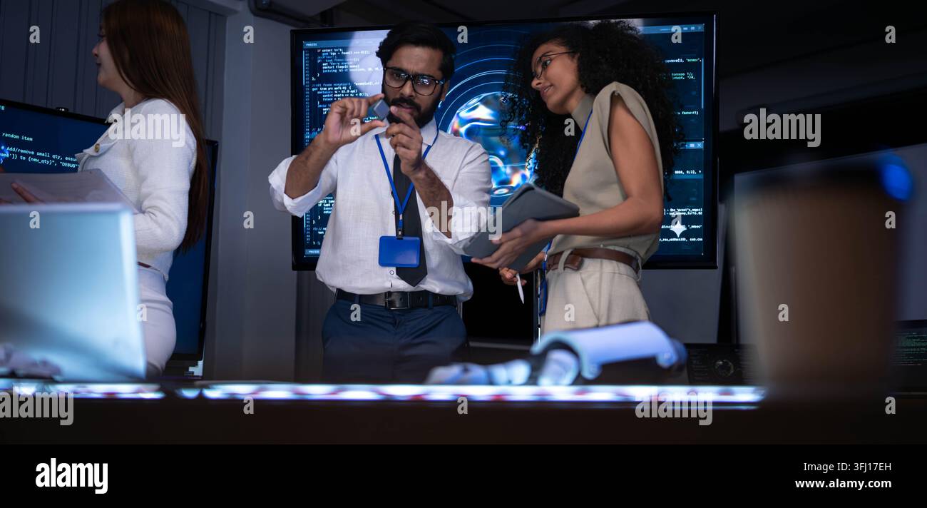 Technology specialists examining electronic component and discussing software engineering solutions in futuristic office with coding screens and digit Stock Photo