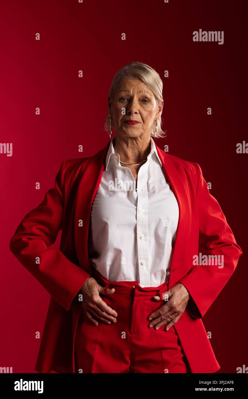 Senior woman posing in studio with red backdrop wearing red suit white shirt gold necklace Stock Photo