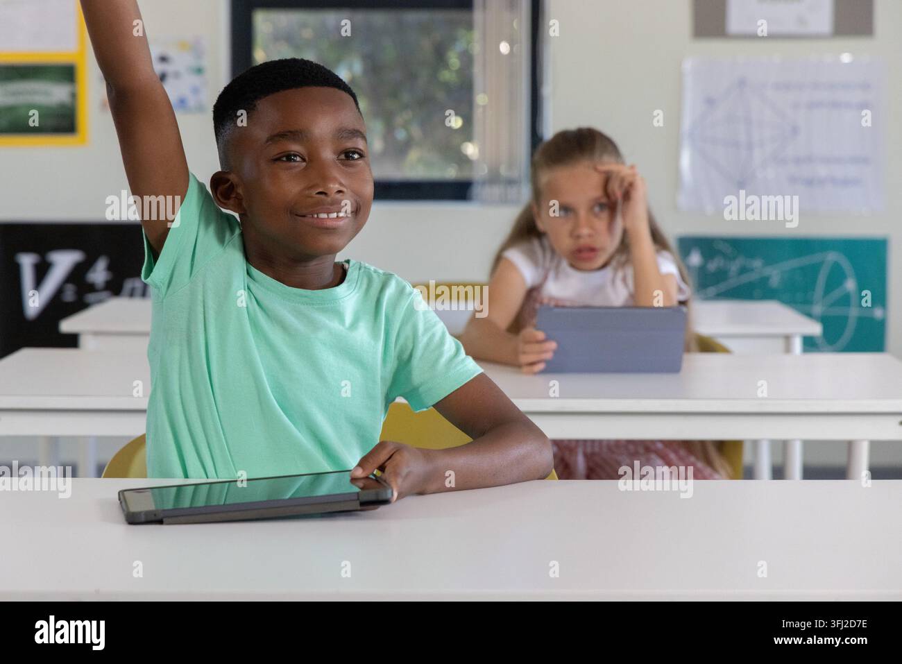 Diverse child classmates raising hand and using tablets in classroom at white desks. Edtech, devices, students, learning, focus, Stock Photo