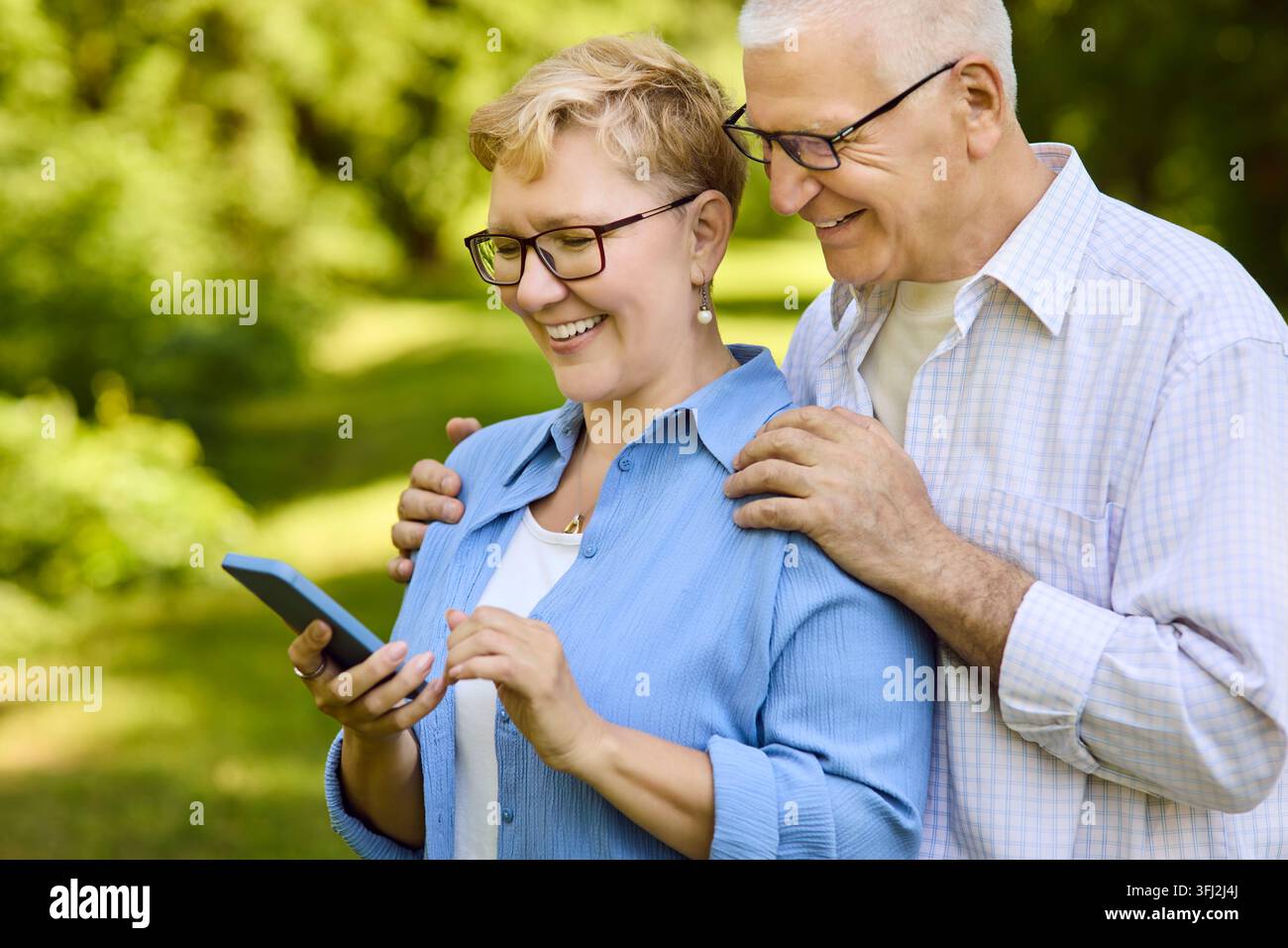 Senior couple laughing using smartphone in park, learning technology outdoors Stock Photo