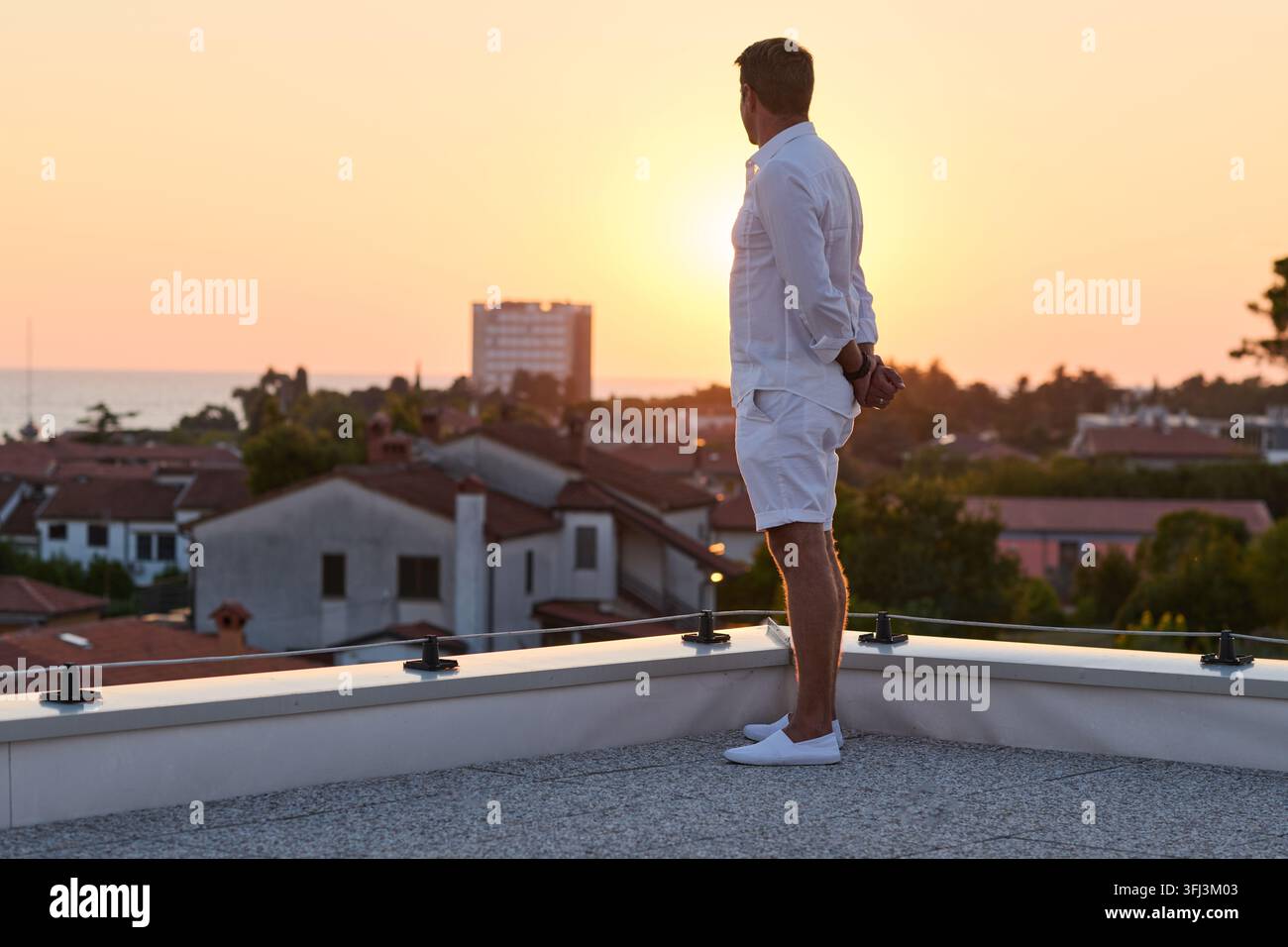 Man Standing on Rooftop Terrace Watching Sunset Over Seaside Town From Home Balcony Stock Photo