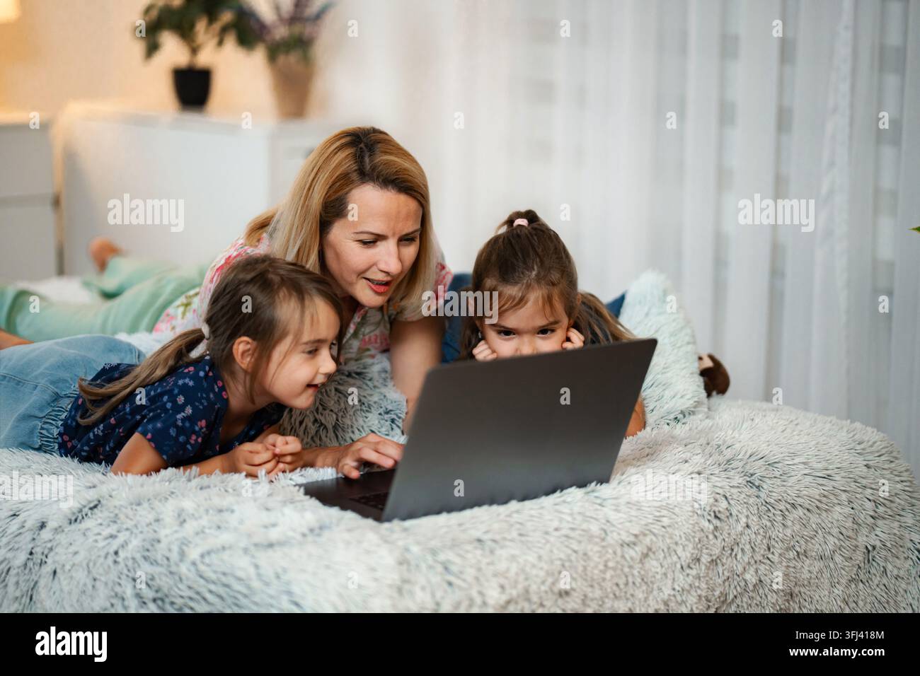 Mother and two daughters lying on bed using laptop for online learning Stock Photo