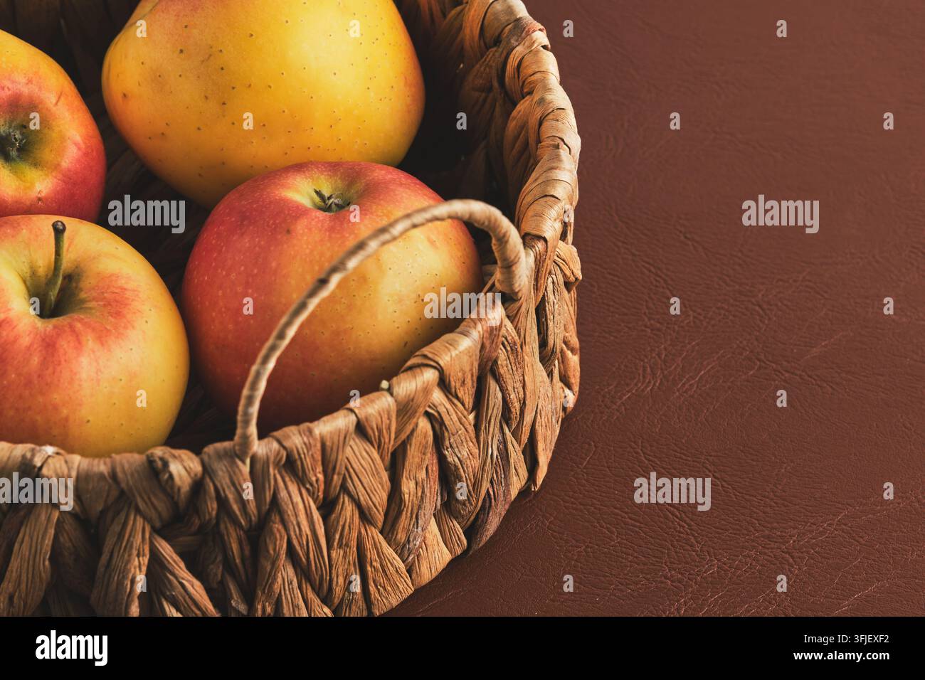 Fresh ripe apples in woven basket on brown leather surface, close up view Stock Photo