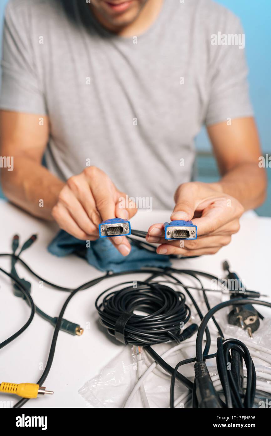 Vertical cropped shot of man holding two VGA connectors, preparing collection of cables, cords, electronic accessories on table. Concept of process of Stock Photo