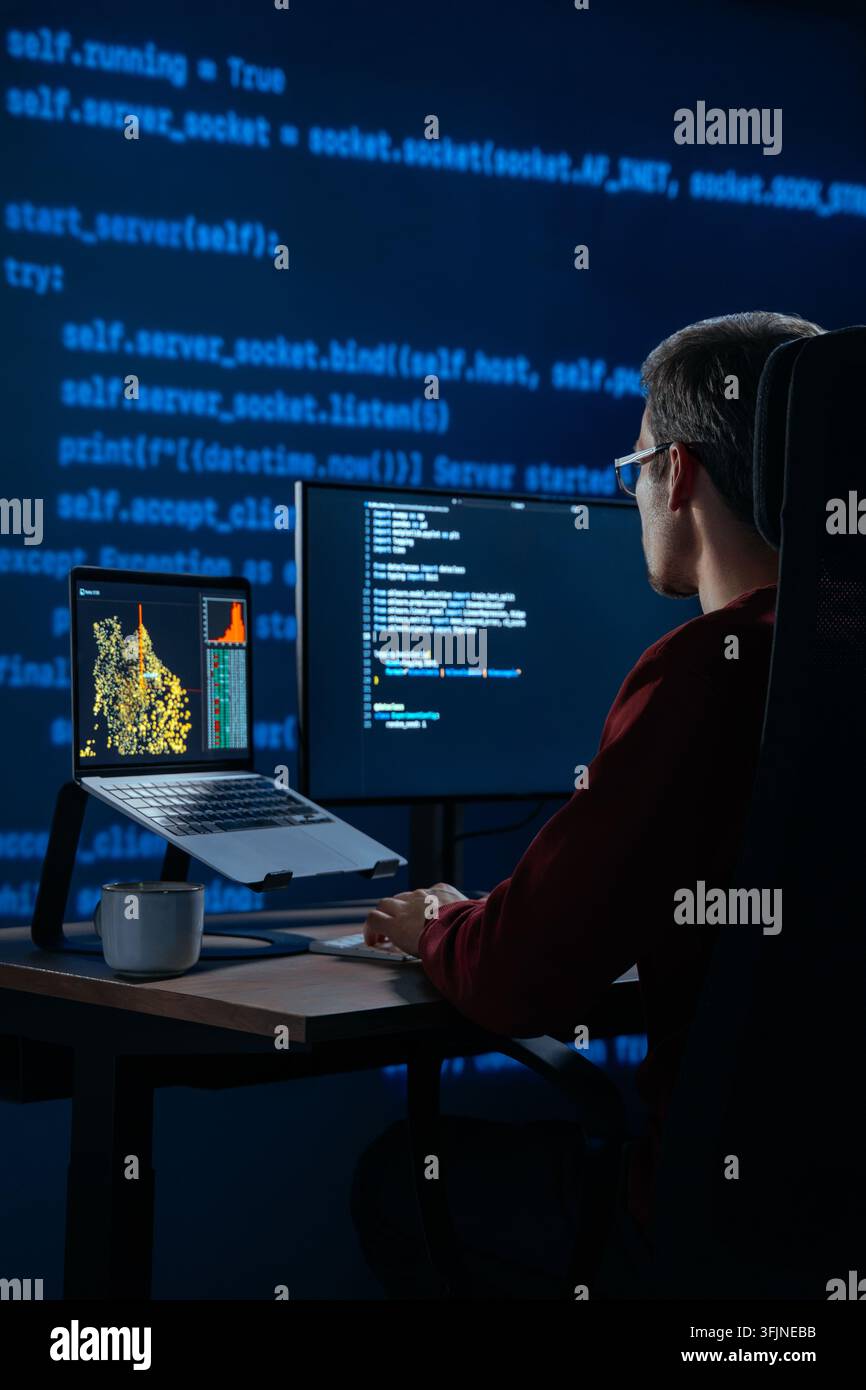 Focused coder works at desk in a dark office with programming code lines running behind. Developer is deeply engaged in a late night data science or AI coding session. Copy space.  Stock Photo