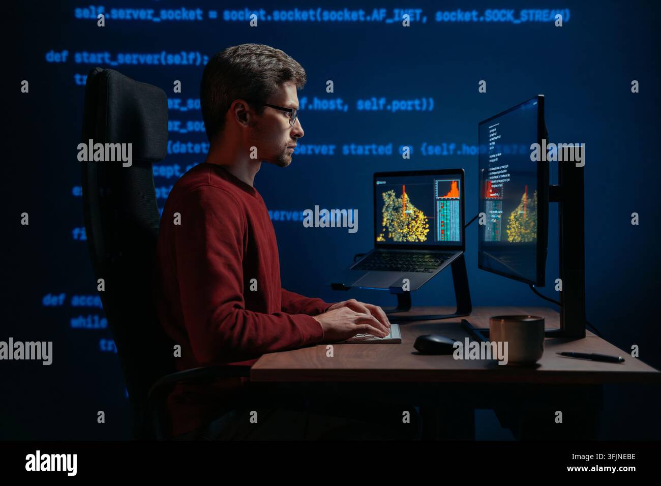Side view, coder at a dark office desk works across multiple screens at night, with programming script on a background. Data scientist working on a machine learning or artificial intelligence task. Stock Photo