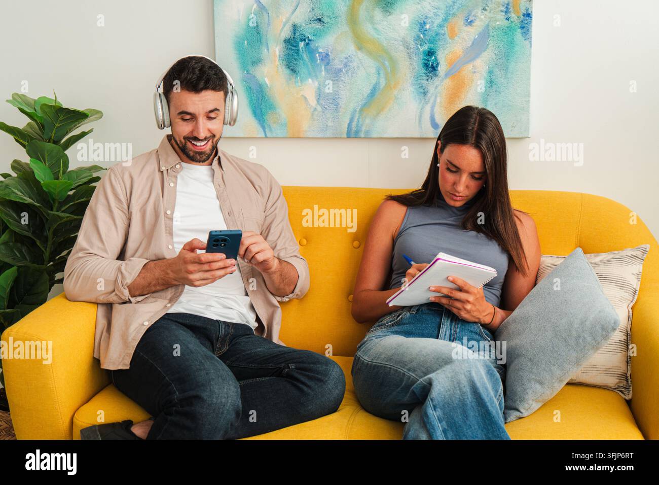 A relaxed couple sits on a bright sofa using a smartphone and writing in a notebook, conveying casual collaboration and shared focus in a comfortable Stock Photo