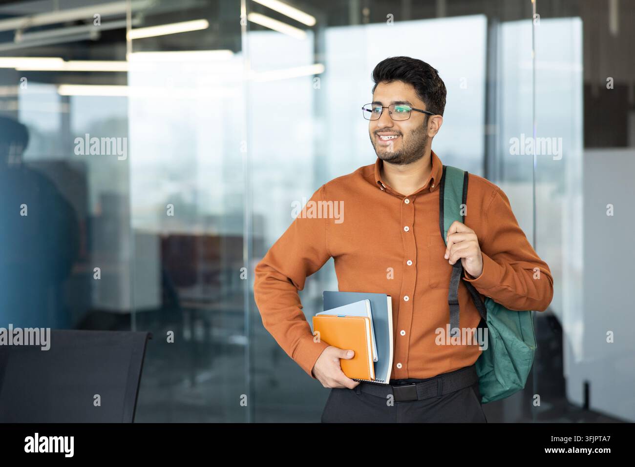 Young professional man with backpack and documents, smiling businessman in modern office environment for corporate career development and workplace success concept. Confident employee holding folders. Stock Photo