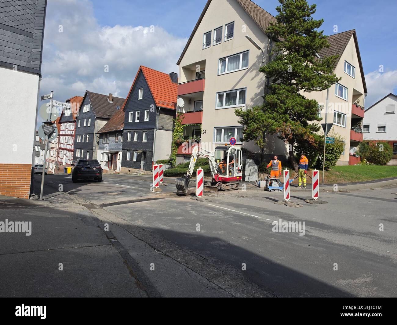 ​Road construction and repair works featuring a small excavator, warning cones, and workers on a street lined with residential buildings - Smartphone Captured Stock Image