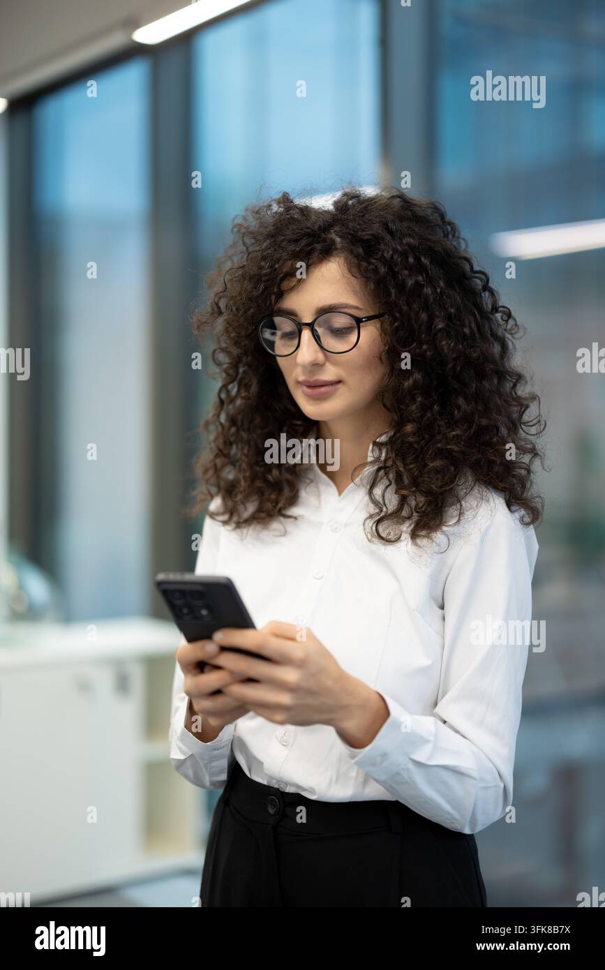 Business communication and digital connectivity concept, young woman using a smartphone for professional tasks in a modern office. Professional employee checking her phone for corporate updates. Stock Photo