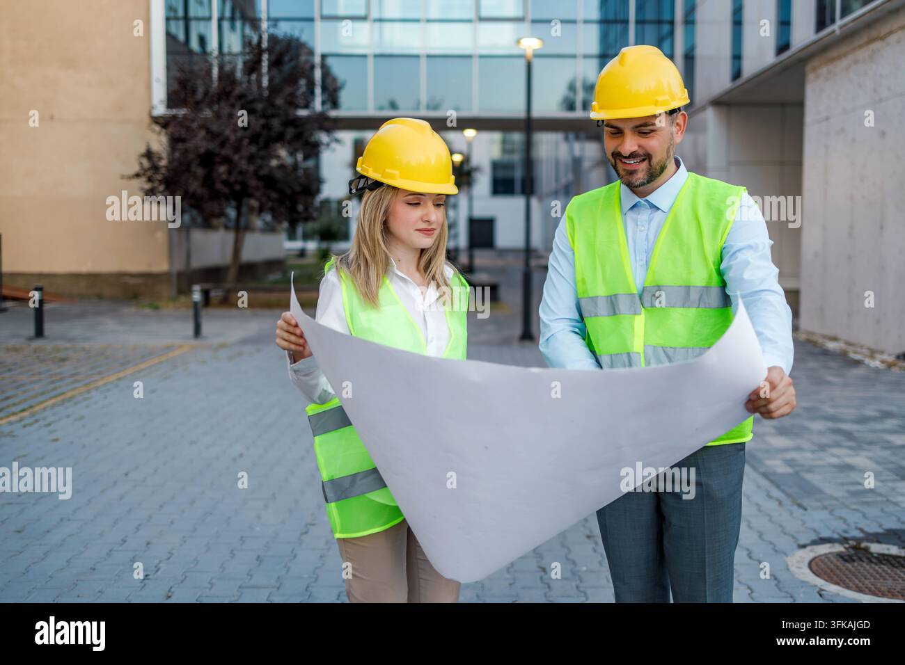 Two construction workers stand together outside a building. They look at plans and discuss their project. Stock Photo