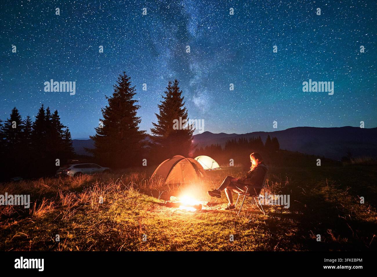 Hiker woman relaxes by campfire, illuminated by its warm glow, with tent nearby under stunning, star-filled sky. Milky Way stretches overhead, adding Stock Photo