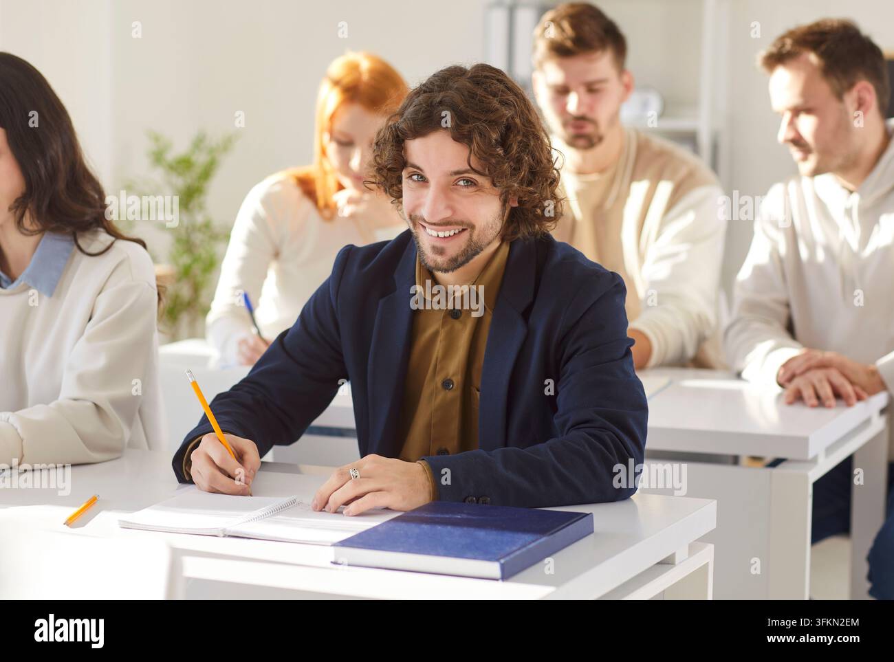 Cheerful curly haired male student writing down notes while having lesson in class Stock Photo