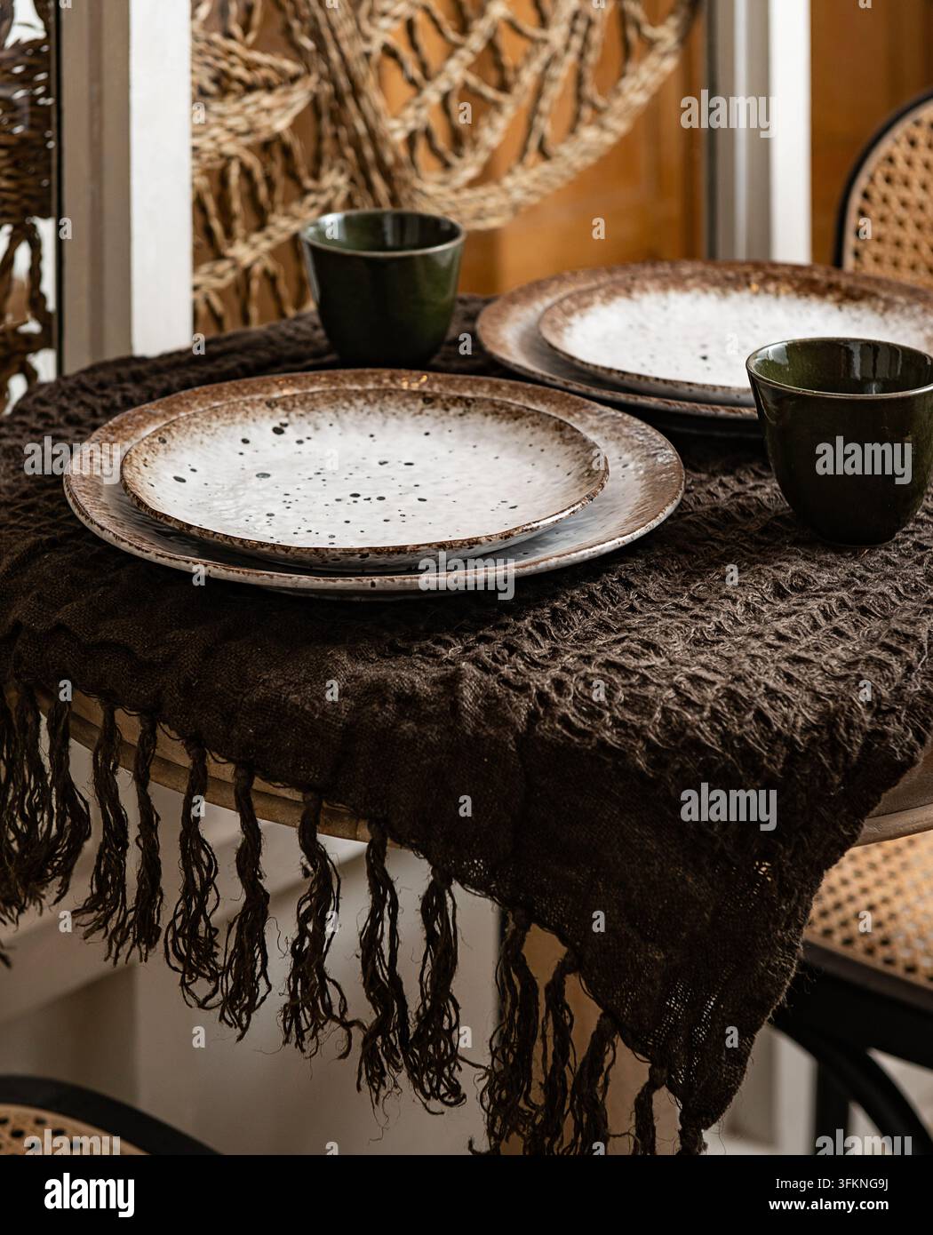 Rustic table setting with stacked speckled ceramic plates, dark green cups, and a fringed dark woven runner. Stock Photo
