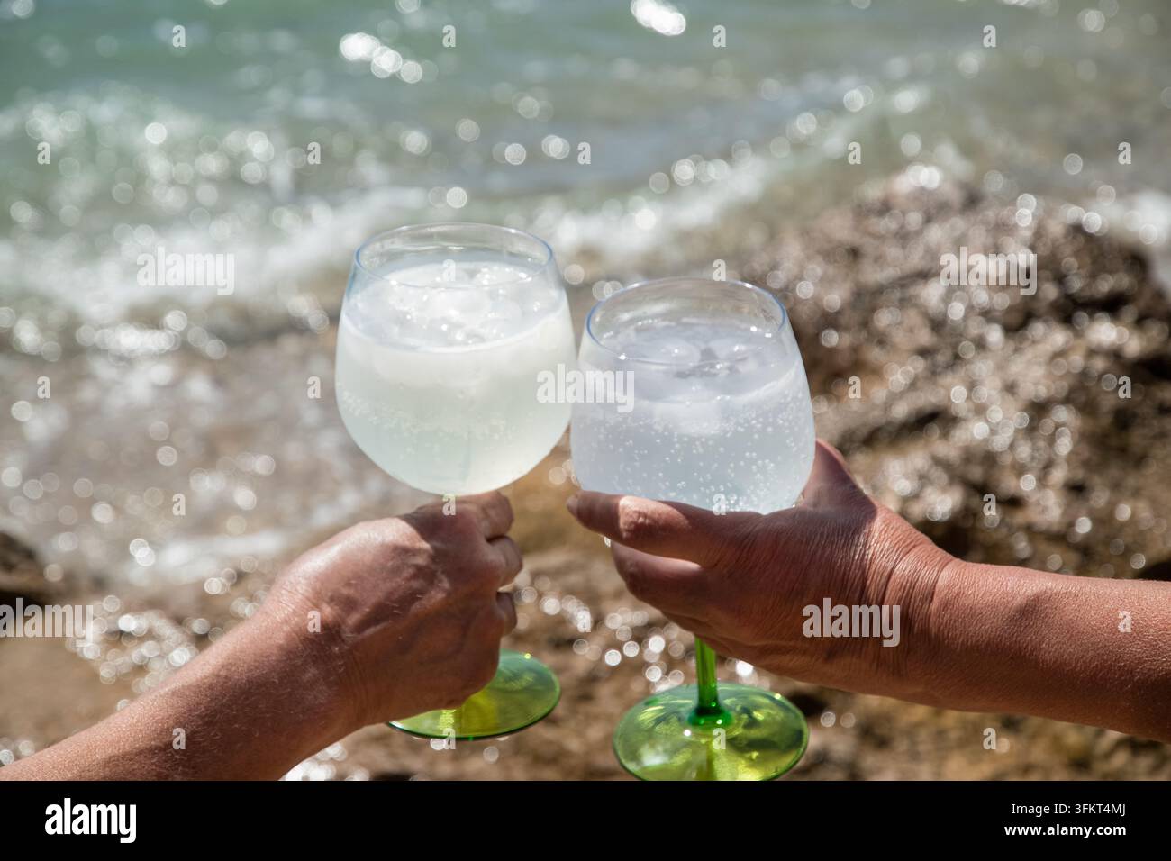 Two people holding glasses of chilled sparkling drinks with ice during a sunny summer day by the sea. Stock Photo
