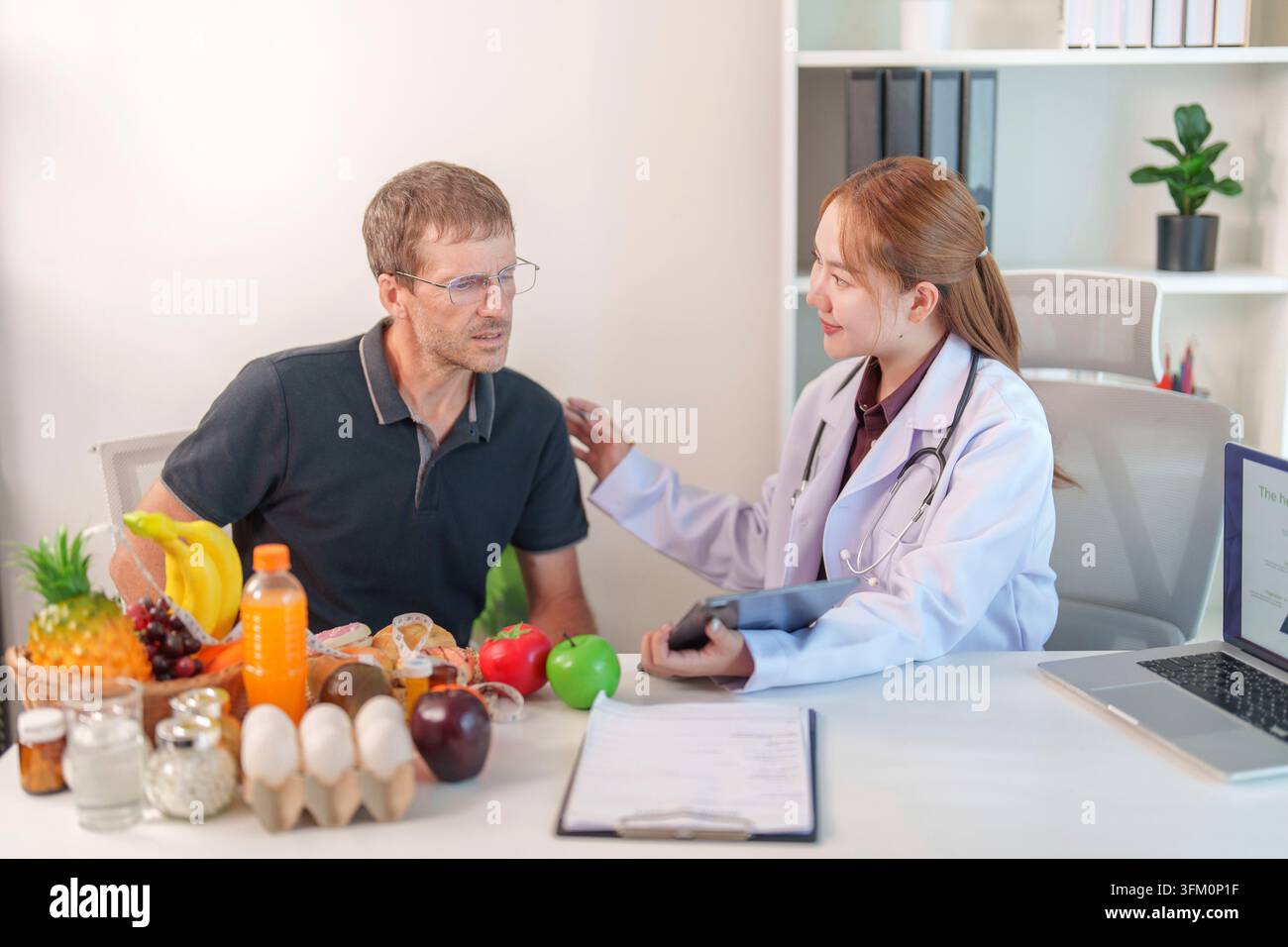Asian female nutritionist with a tablet, calming down male patient sitting on clinic desk filled with healthy food options Stock Photo