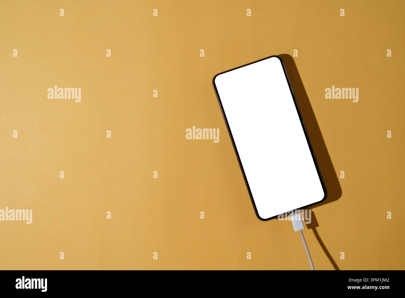 An overhead flat lay of a smartphone with a blank white screen plugged into a white charging cable on a solid yellow background Stock Photo