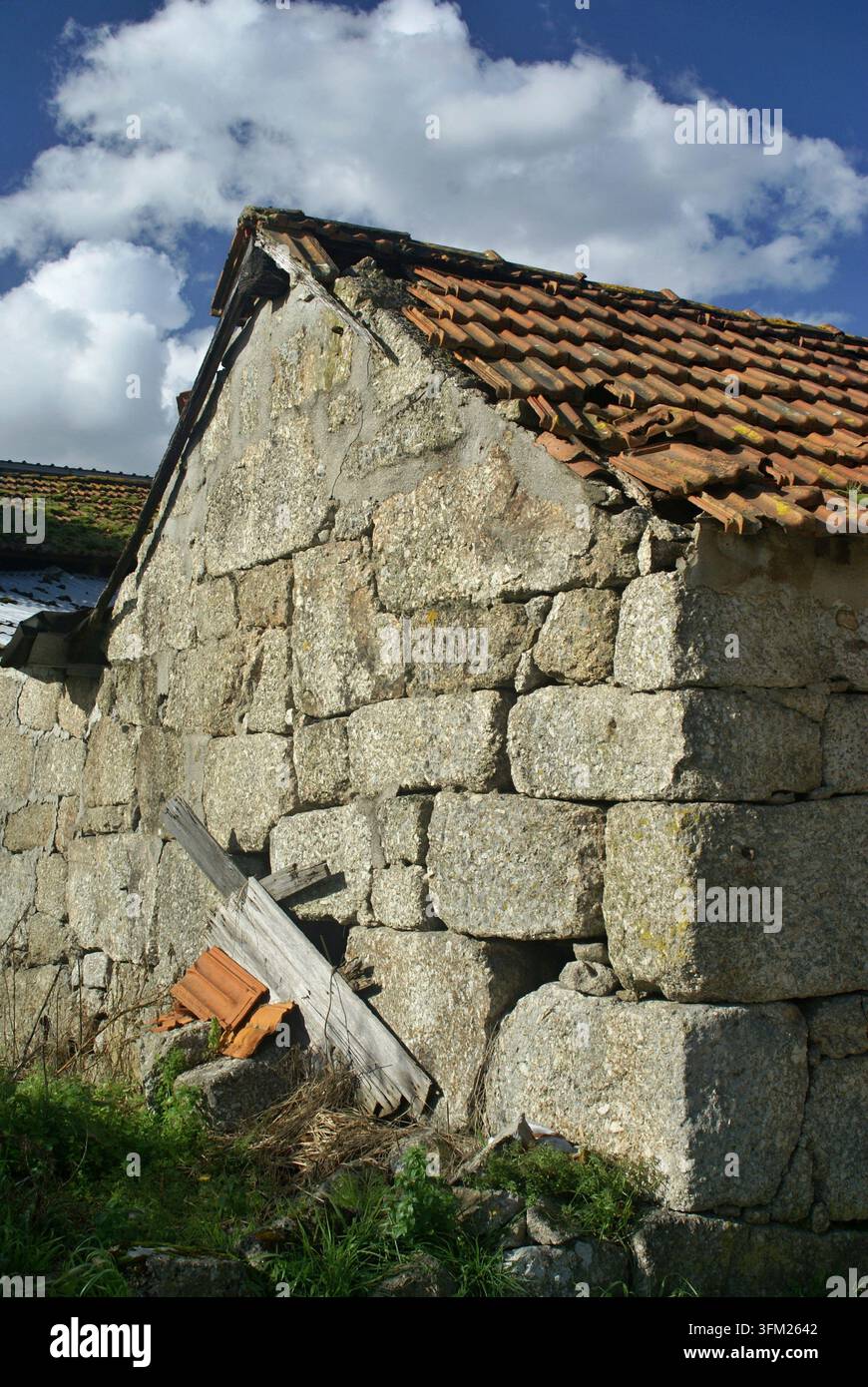Weathered stone house wall with a broken tile roof under a blue sky Stock Photo
