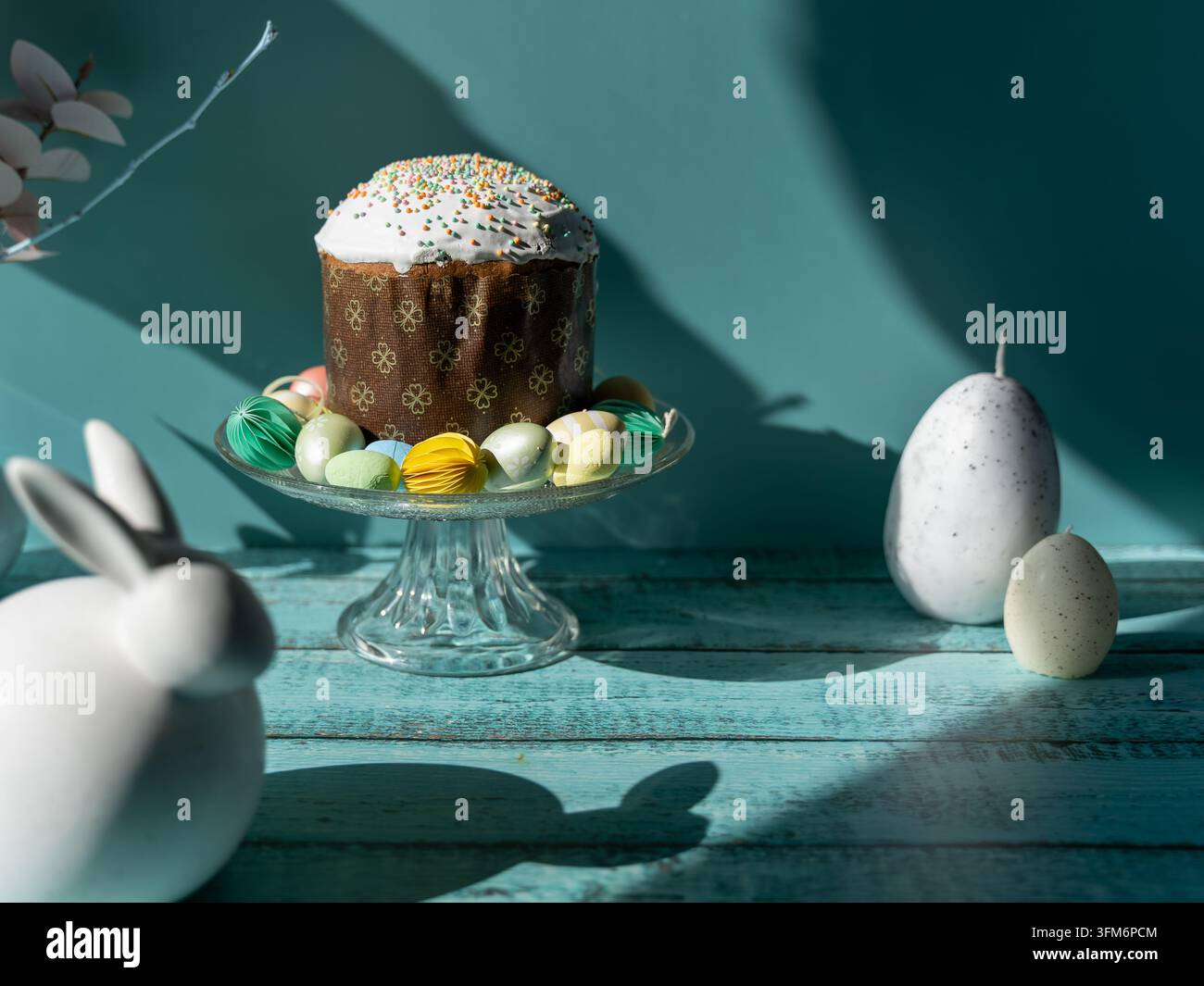 Traditional Paska Pasca Easter bread served on a glass cake stand with decorative Easter eggs and a white bunny ornament on a turquoise wooden backgro Stock Photo