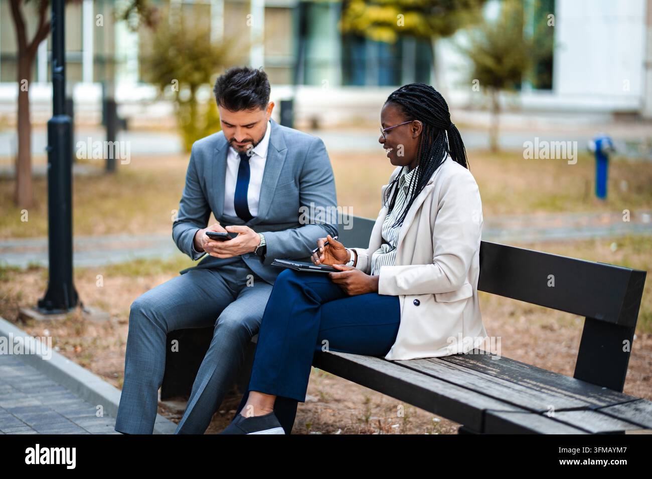 Diverse business professionals sharing ideas and using digital devices while sitting on a park bench Stock Photo