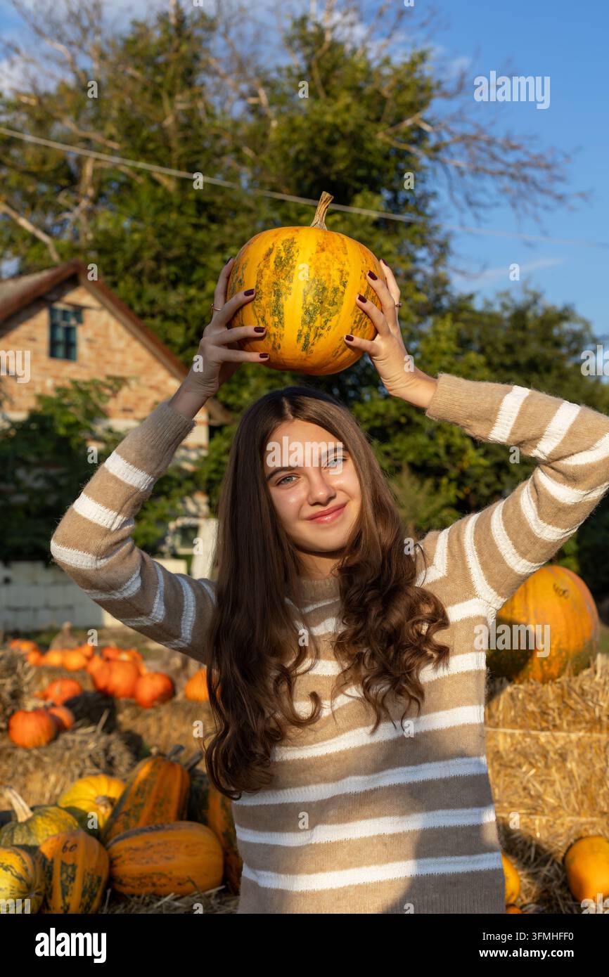 Young woman holding a yellow pumpkin above her head in a sunny autumn garden, cheerful harvest portrait surrounded by hay bales and colorful pumpkins. Stock Photo
