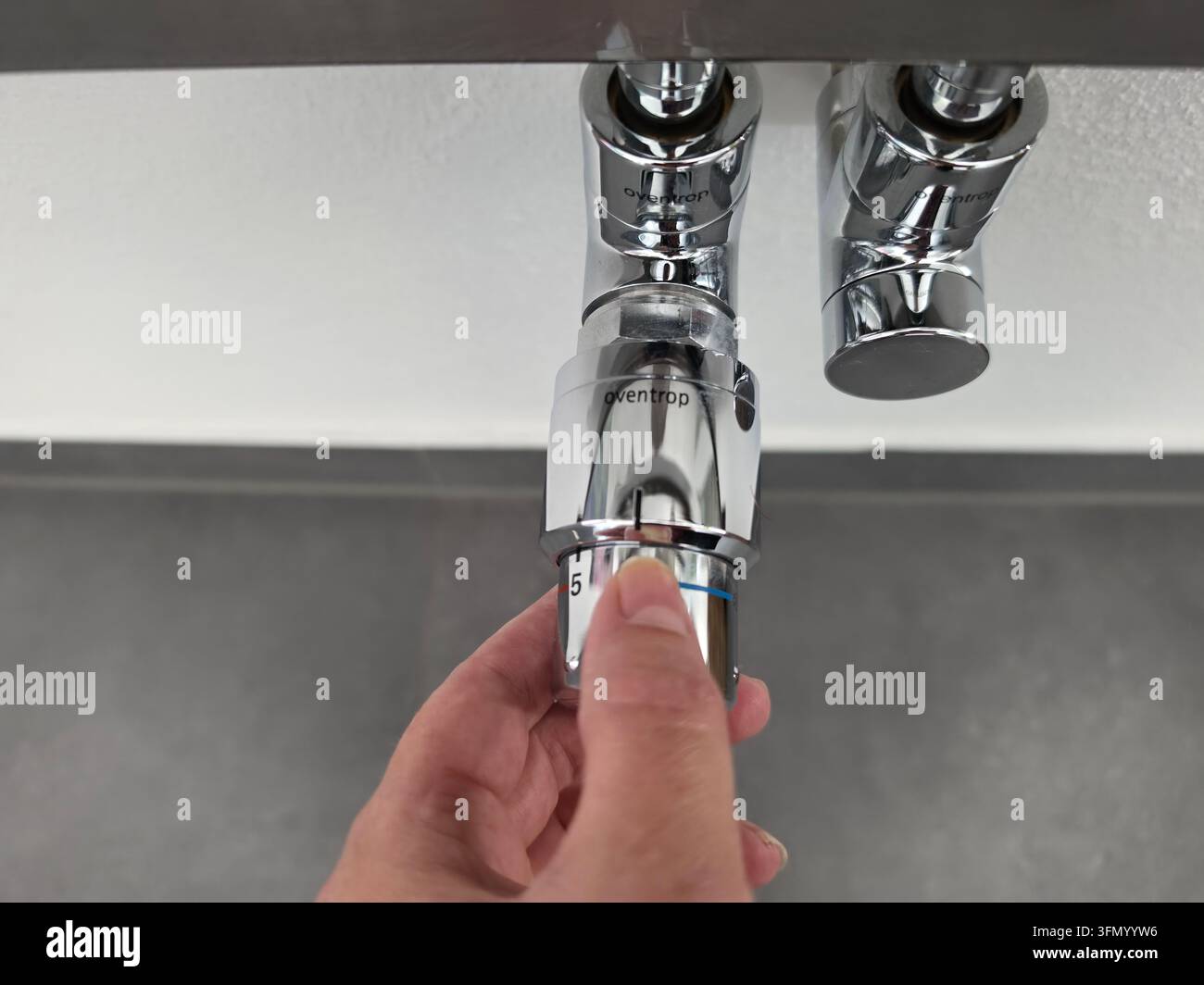 Person turning thermostatic radiator valve on chrome heating pipes mounted on grey wall, close-up view of manual temperature adjustment in home interi Stock Photo
