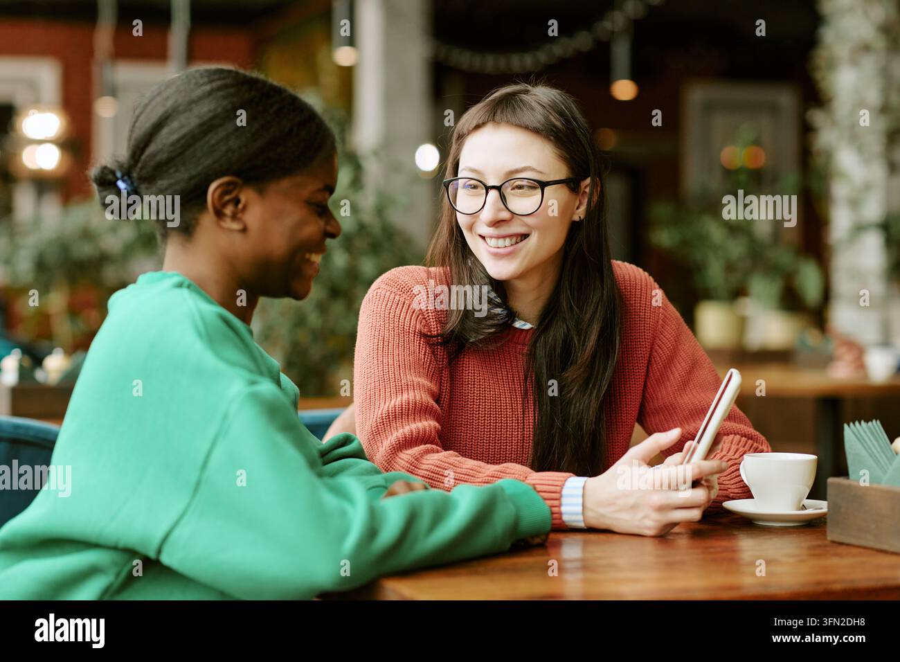 Diverse Female Friends Talking at Cafe Table Using Smartphone Stock Photo
